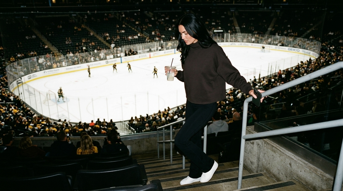 What to wear to a hockey game for women: a woman in a dark espresso Rhone DreamGlow mock neck and black Rhone Revive flare leggings walking down the arena section steps with a drink as players warm up on the ice below