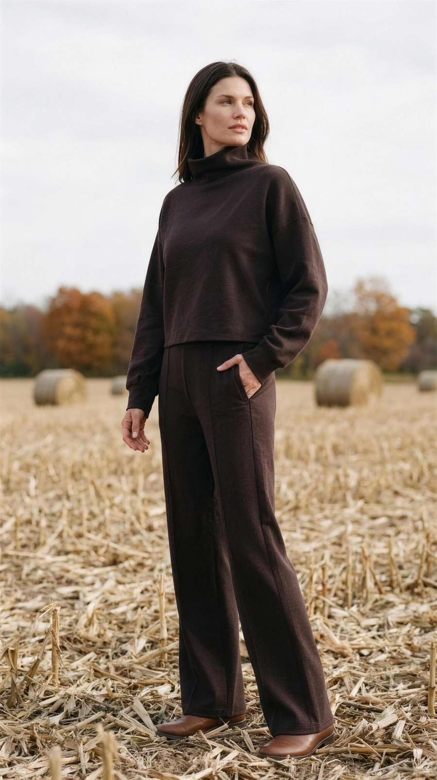 DreamGlow Mock Neck and Trouser - Dark Espresso — Door County: the matching espresso set, boxy funnel-neck knit over the wide-leg trouser with its pressed crease, standing in pale corn stubble with round bales beyond
