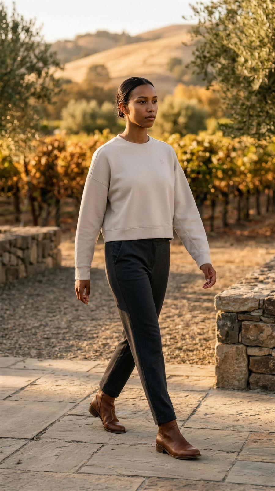 Rhone DreamGlow Crew Neck in Sand, cropped and boxy over dark charcoal trousers, in a stone vineyard courtyard in Napa Valley at golden hour in October.